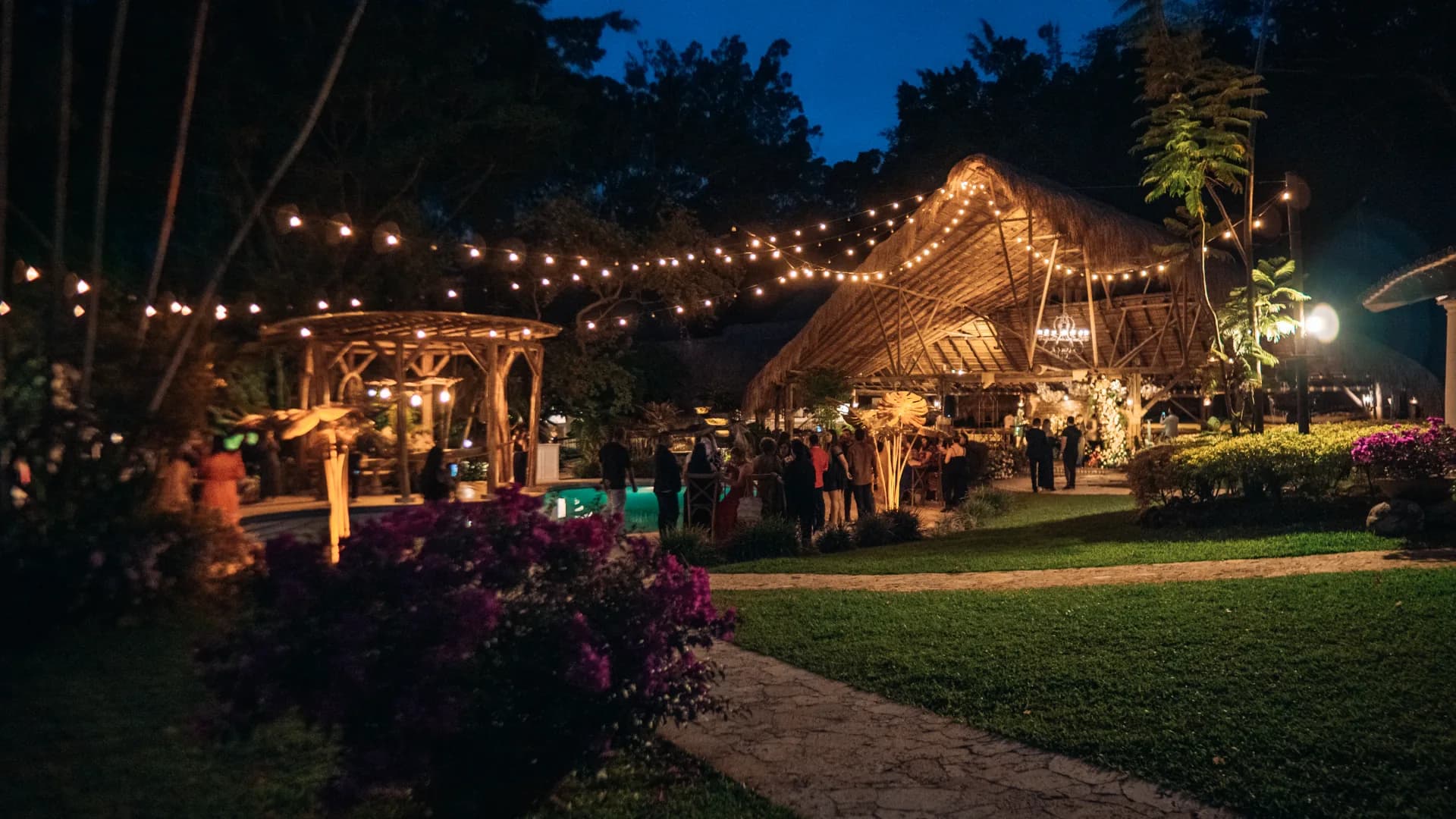 Guests with drinks at the poolside cocktail area, under string lights and a palm-thatched roof at dusk