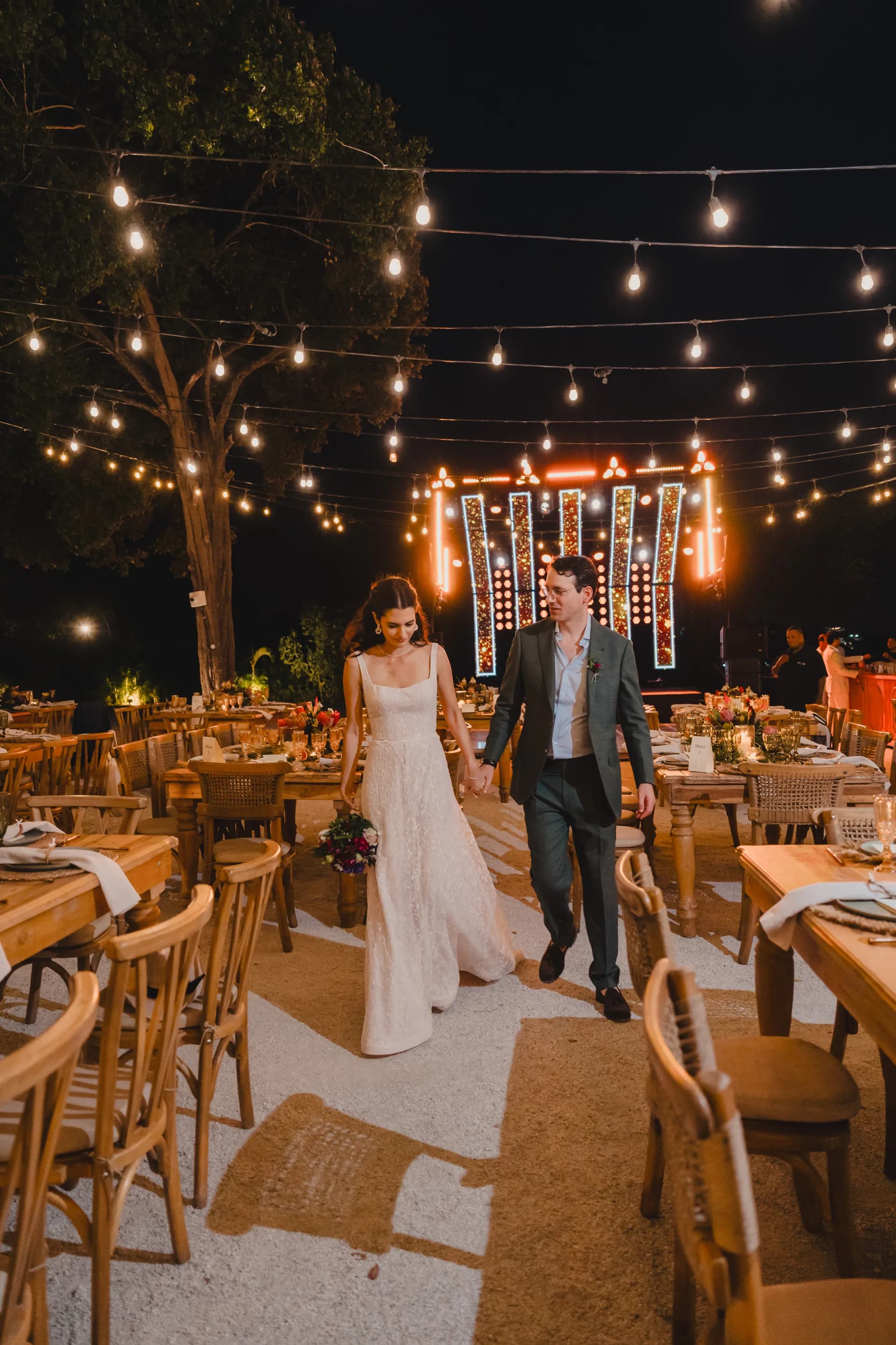 The couple walking hand in hand through the reception at night, under string lights